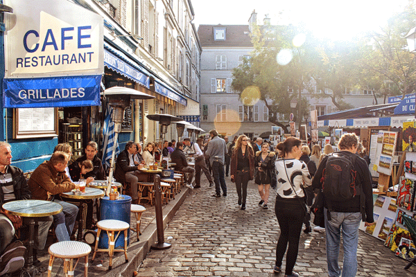Lots of people sitting at tables outside a Paris cafe and people walking in a cobbled street on a sunny day